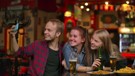 group-of-friends-man-and-two-girls-take-selfie-in-a-bar-laughing-and-smiling.-Spending-time-with-dumbfounds-in-a-bar